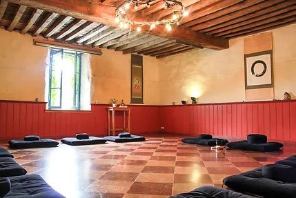 een rustig ingerichte ruimte met warme kleuren, langs de muren liggen meditatiematten en kussens, links is een raam, rechts een scroll met enso